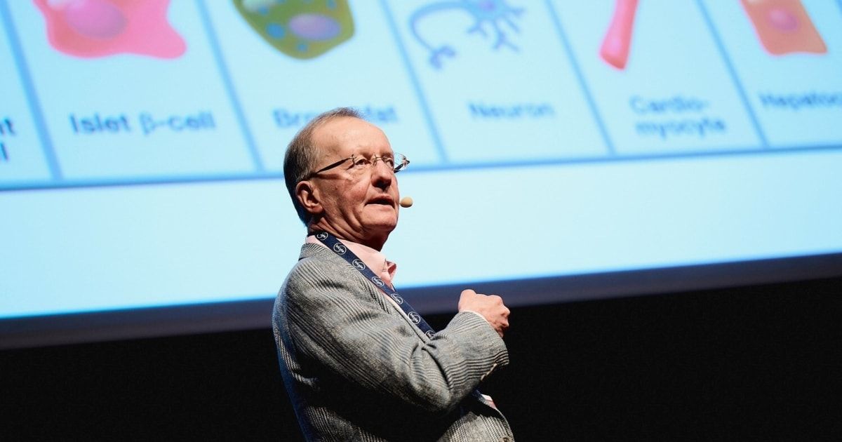 Ein älterer Mann mit Brille hält eine Rede. Hinter ihm zeigt eine Leinwand Bilder von Zellen und Neuronen.