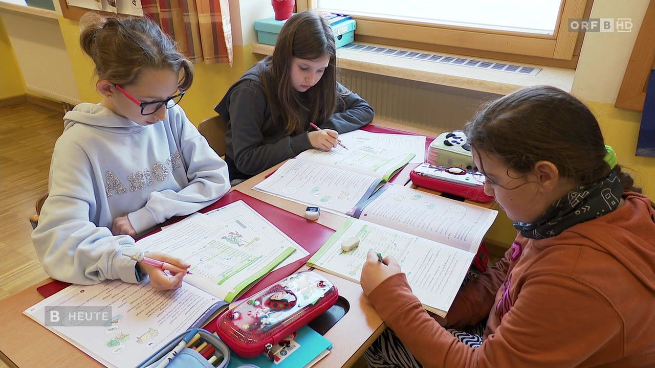 Drei Mädchen sitzen an einem Schreibtisch und schreiben in ihre Notizbücher. Zwei von ihnen tragen Brillen. Offene Bücher, ein Federmäppchen und andere Schulsachen liegen auf dem Tisch.