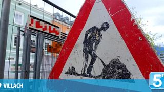 Ein rotes und weißes dreieckiges Schild mit einer graben bauenden Figur. Es befindet sich in der Nähe eines Gebäudes und einer Metallumzäunung.