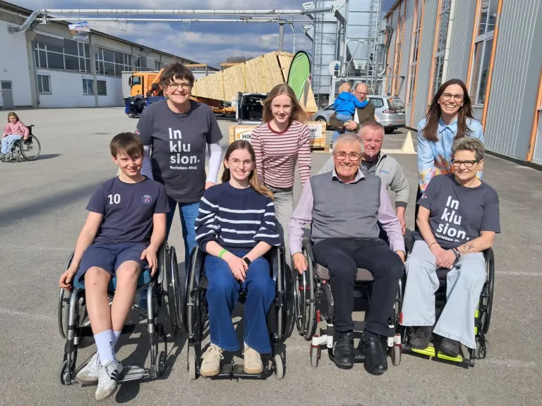 Eine Gruppe von Menschen im Rollstuhl, die für ein Foto in einer Außenumgebung lächeln. Dahinter hält eine Person ein Kind. Im Hintergrund sind verschiedene Fahrzeuge und ein Gebäude zu sehen.