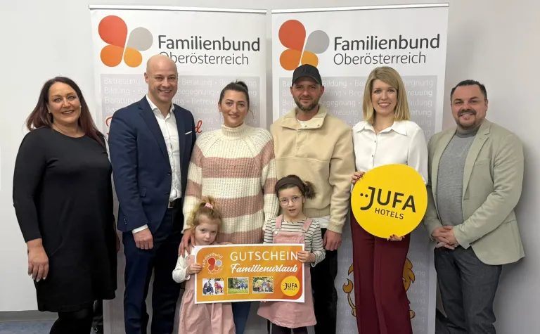 Mehrere Erwachsene und Kinder stehen lächelnd vor einem Banner. Sie halten ein Schild und einen runden Geschenkgutschein von JUFA Hotels. Das Banner im Hintergrund hat Text auf Deutsch.
