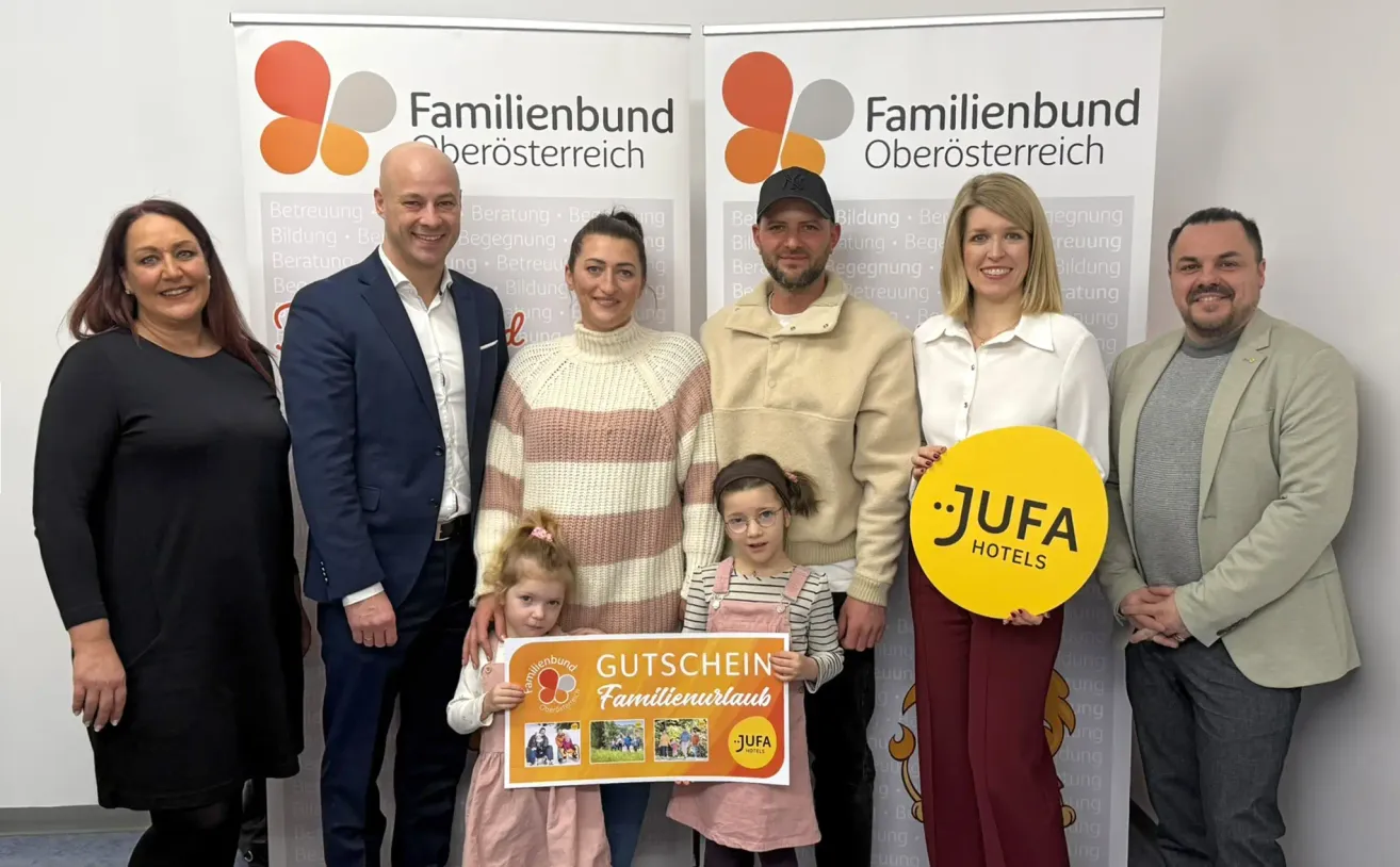 Mehrere Erwachsene und Kinder stehen lächelnd vor einem Banner. Sie halten ein Schild und einen runden Geschenkgutschein von JUFA Hotels. Das Banner im Hintergrund hat Text auf Deutsch.
