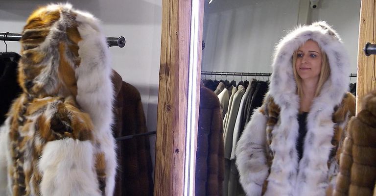 Eine Frau in einem Pelzmantel steht vor einem Spiegel. Hinter ihr sind Pelzmäntel an Kleiderbügeln ausgestellt. Die Pelzmäntel sind weiß, braun und schwarz.