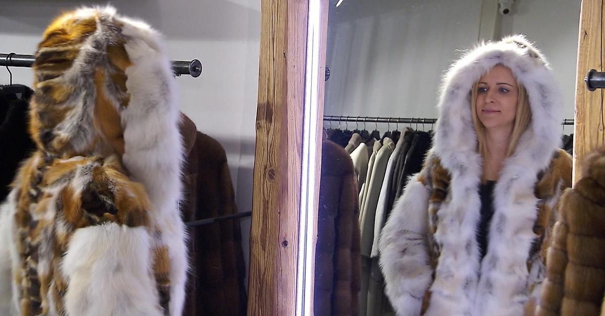 Eine Frau in einem Pelzmantel steht vor einem Spiegel. Hinter ihr sind Pelzmäntel an Kleiderbügeln ausgestellt. Die Pelzmäntel sind weiß, braun und schwarz.