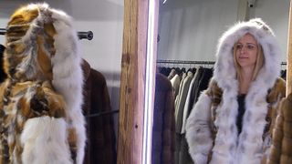 Eine Frau in einem Pelzmantel steht vor einem Spiegel. Hinter ihr sind Pelzmäntel an Kleiderbügeln ausgestellt. Die Pelzmäntel sind weiß, braun und schwarz.