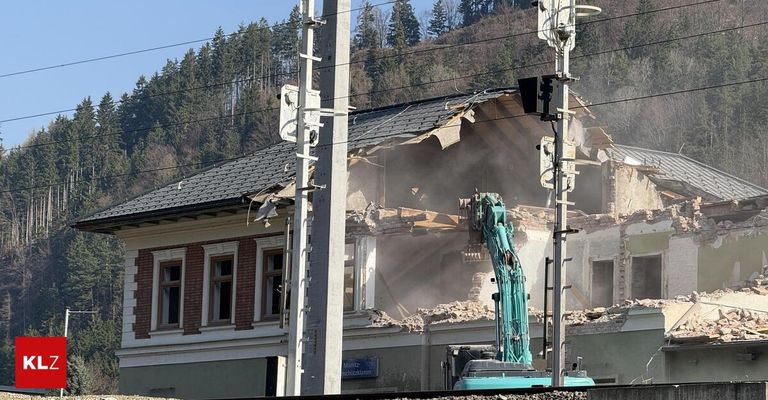 Ein Abrissgelände mit einem großen Gebäude im Abrisszustand mit einem grünen Bagger in der Nähe eines Strommastes und einem Schild am Gebäude.