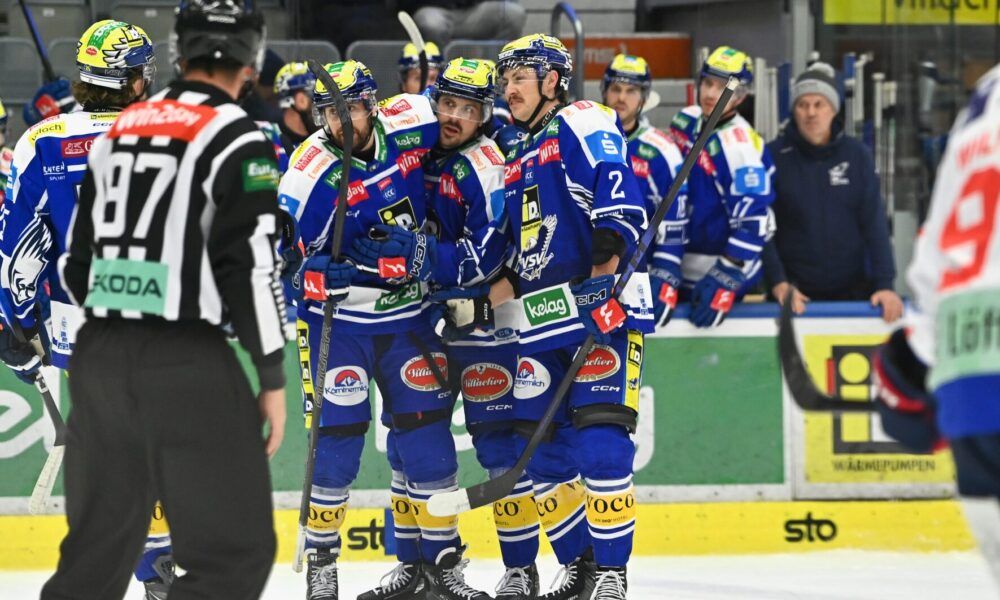 Eishockeyspieler in blauen Uniformen feiern auf dem Eis. Ein Schiedsrichter steht in der Nähe, und Zuschauer beobachten von den Tribünen aus. Die Eisbahn hat verschiedene Werbungen an ihren Barrieren.