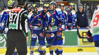 Eishockeyspieler in blauen Uniformen feiern auf dem Eis. Ein Schiedsrichter steht in der Nähe, und Zuschauer beobachten von den Tribünen aus. Die Eisbahn hat verschiedene Werbungen an ihren Barrieren.