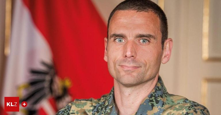 Ein lächelnder Mann in einer Camouflage-Uniform steht vor einer roten Flagge mit einem Symbol.