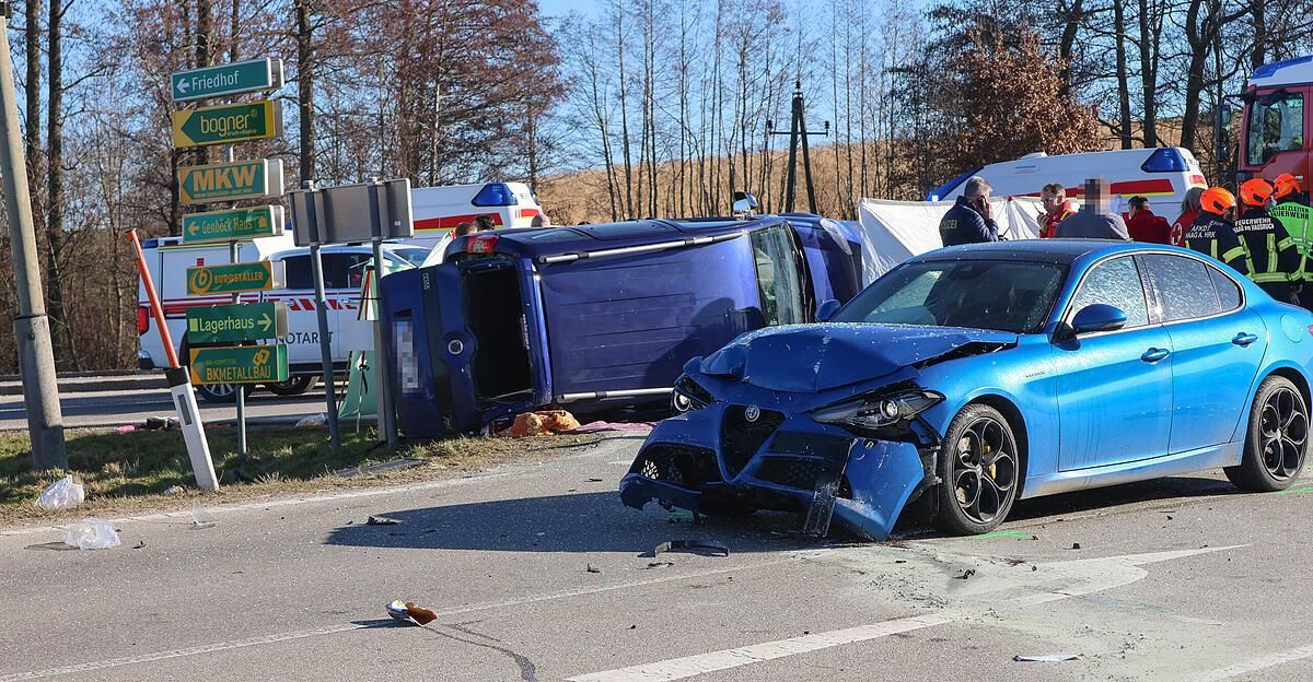 Ein blaues Auto ist auf dem Kopf auf der Straße. Ein anderes Auto hat einen beschädigten Front. Es stehen Leute herum, und Rettungsfahrzeuge sind in der Nähe.