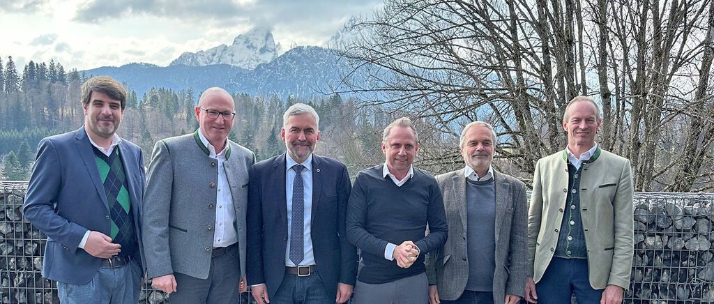 Fünf Männer in formeller Kleidung stehen lächelnd im Freien, mit Bergen und Bäumen im Hintergrund.