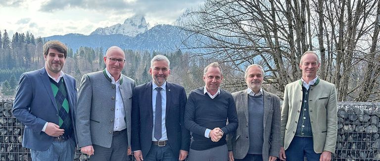 Fünf Männer in formeller Kleidung stehen lächelnd im Freien, mit Bergen und Bäumen im Hintergrund.