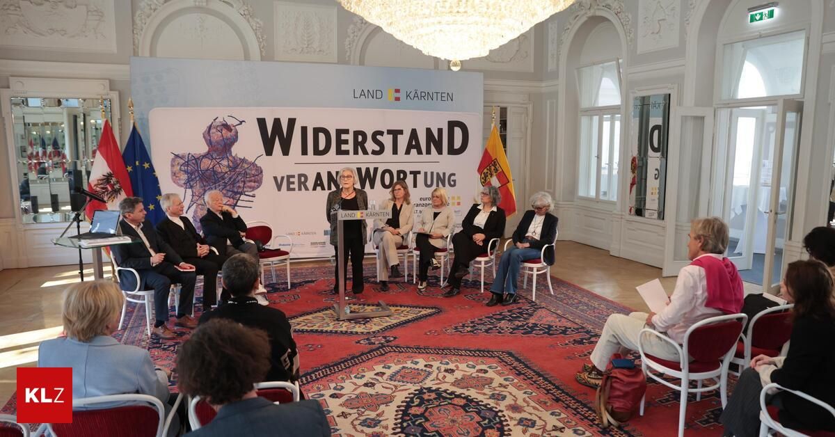 Eine Veranstaltung in einem großen Saal mit einem großen Schild, das 'Widerstand' steht und einer Frau, die an einem Podium spricht. Die Menschen sitzen auf Stühlen um einen roten Teppich. Eine Flagge ist im Hintergrund.