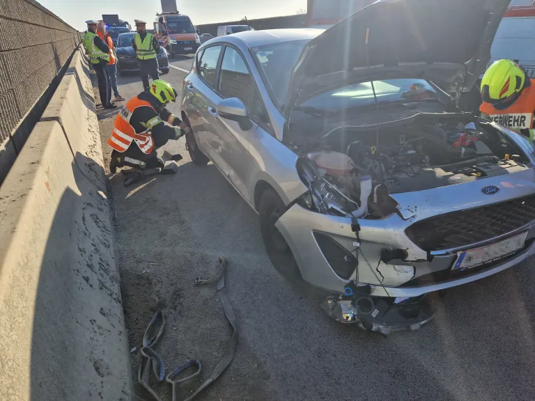 Ein Auto mit offenem Motorraum und beschädigter Front wird von einem Notfallsanitäter am Straßenrand untersucht.