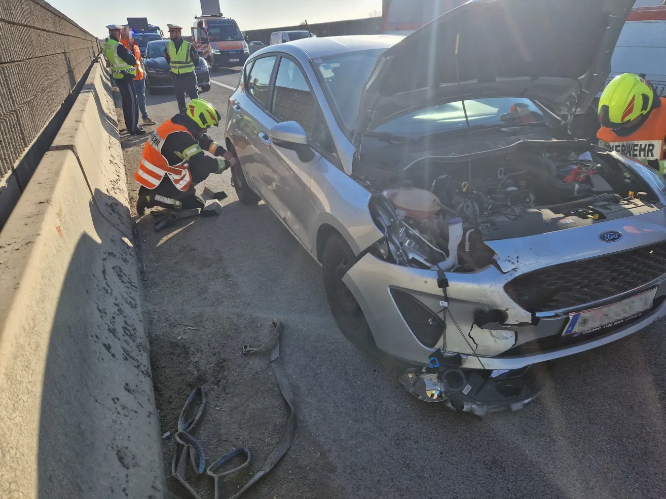 Ein Auto mit offenem Motorraum und beschädigter Front wird von einem Notfallsanitäter am Straßenrand untersucht.