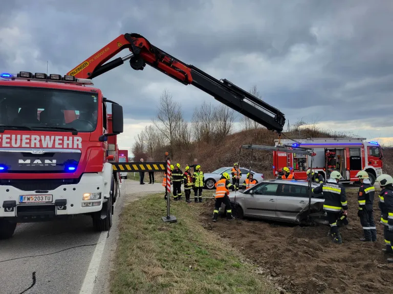 Ein roter Feuerwehrwagen mit einem Kran hebt ein demoliertes silbernes Auto an. Rettungskräfte in orangen Westen und Helmen helfen. Die Szene ist an einer grasigen Straßenseite.