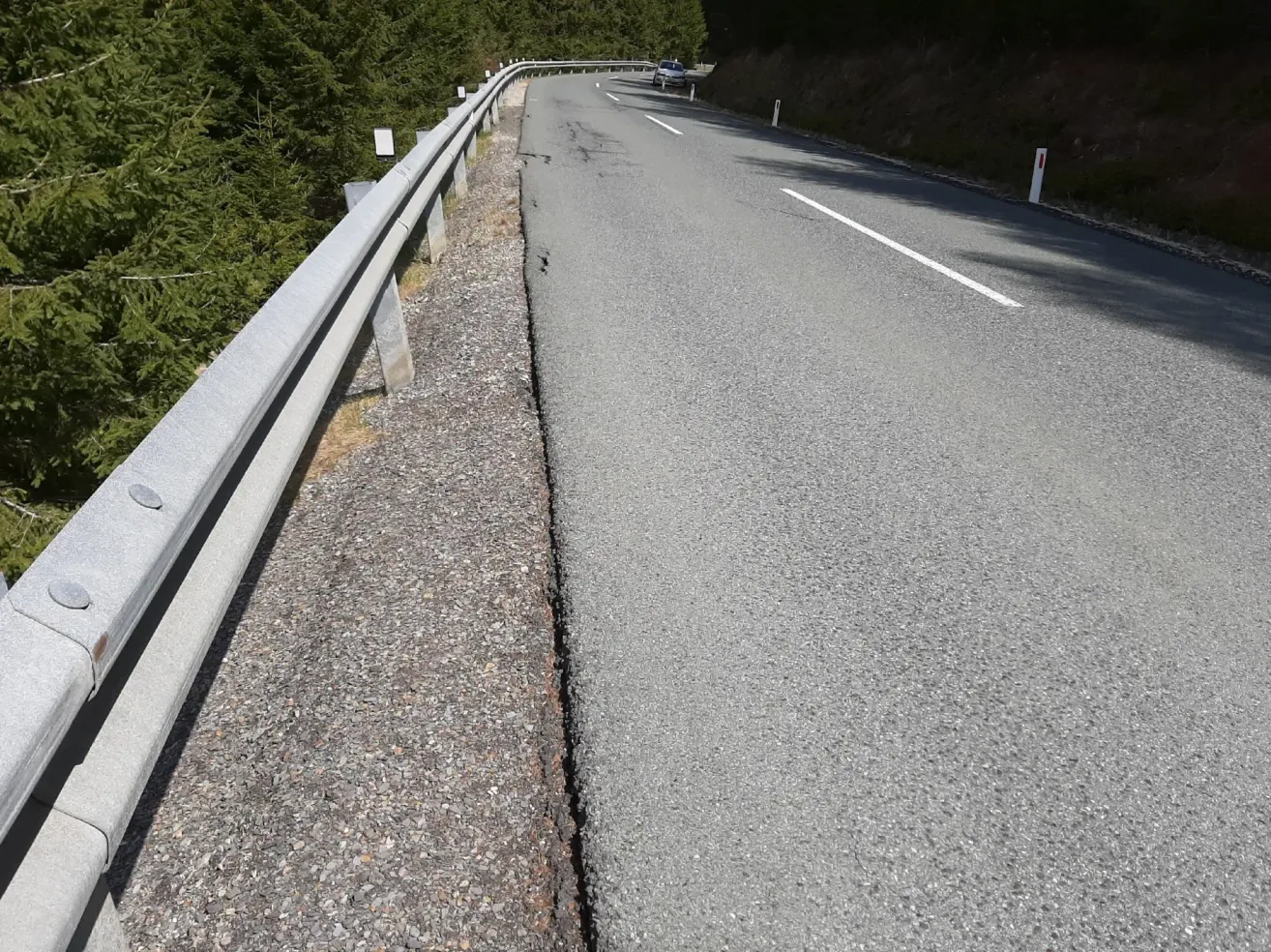 Eine Straße mit beschädigtem Asphalt und einem Auto in der Ferne. Bäume säumen den Straßenrand.
