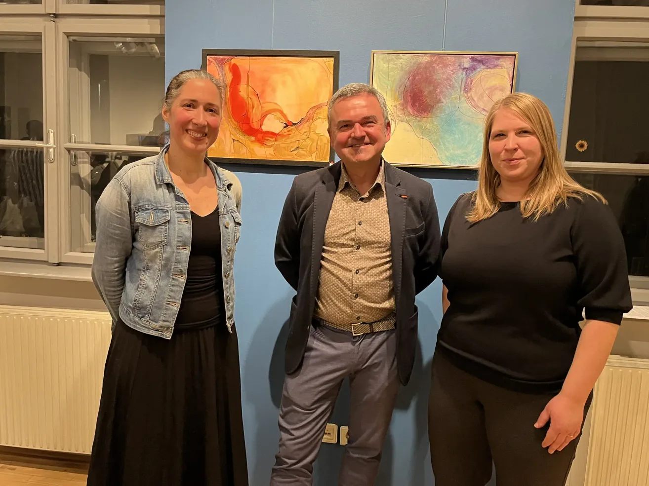 Drei Personen stehen gemeinsam vor einer blauen Wand mit zwei abstrakten Gemälden. Die Frau links trägt eine Jeansjacke und einen schwarzen Rock. Der Mann in der Mitte hat ein braunes Hemd, einen schwarzen Blazer und einen Gürtel. Die Frau rechts hat blonde Haare und trägt ein schwarzes Oberteil.