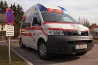 Ein Krankenwagen steht am Straßenrand und zeigt ein rotes Kreuz an seiner Seite. Das Fahrzeug ist ein Volkswagen mit dem Kennzeichen '11'. Hinter dem Krankenwagen ist ein Schild mit einem blau-weißen X zu sehen.