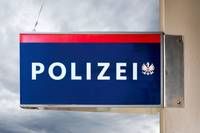 Ein blau-roter Schild mit weisser Schrift, das 'POLIZEI' besagt, hängt an einem Gebäude vor einem bewölkten Himmel.