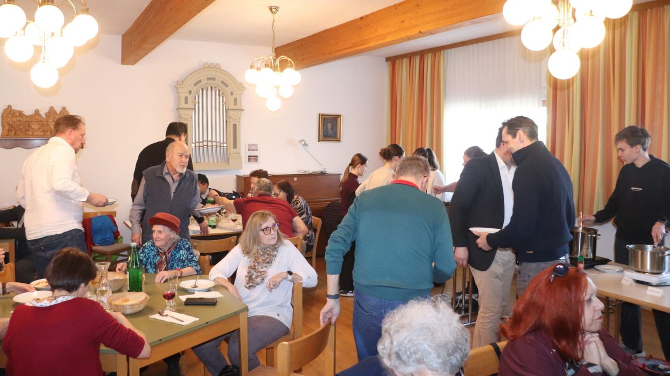A dining room with a piano and chandelier, several people are seated and standing. A man holds a plate and looks around. A woman with glasses is seated with a scarf.