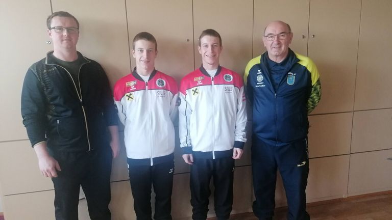 Drei Männer in Sportkleidung stehen zusammen, zwei tragen rote und weiße Jacken mit 'SILUS' darauf und der dritte eine blaue Jacke. Sie lächeln alle und scheinen für ein Foto zu posieren.