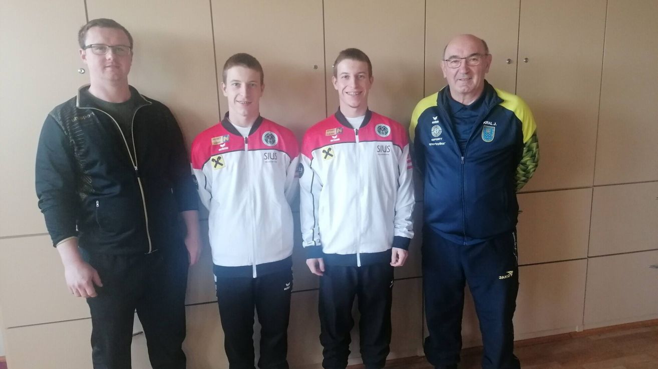 Drei Männer in Sportkleidung stehen zusammen, zwei tragen rote und weiße Jacken mit 'SILUS' darauf und der dritte eine blaue Jacke. Sie lächeln alle und scheinen für ein Foto zu posieren.