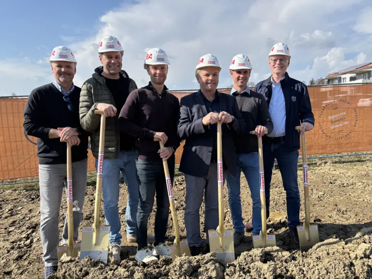 Eine Gruppe von sechs Männern in Schutzhelmen posiert für ein Foto an einer Baustelle und hält Schaufeln. Hinter ihnen befindet sich ein Zaun und ein bewölkter Himmel.