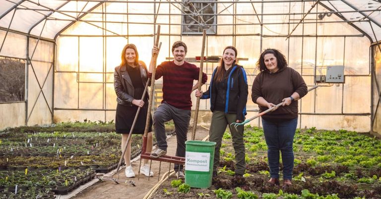 Four people in a greenhouse, three women and one man, smiling, with gardening tools and a compost bin, standing on soil with plants and seedlings.