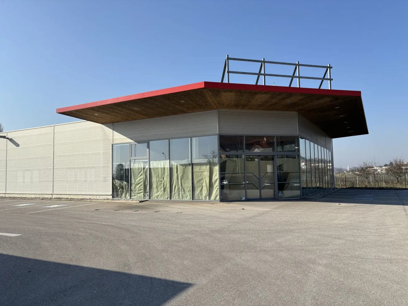 A modern building with a red roof and large glass windows stands in an empty lot under a clear blue sky.