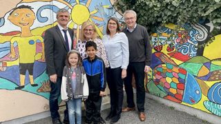 A family of five, including two adults and three children, stands together in front of a colorful mural. They all wear formal attire.