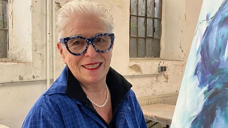 An elderly woman with white hair is smiling and wearing glasses. She is dressed in a blue jacket and a pearl necklace. Behind her is a window with a damaged frame.