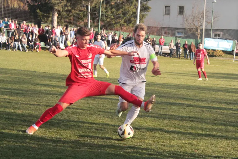 Zwei Fußballspieler sind auf einem Feld, einer in Rot und der andere in Weiß. Der rote Spieler tritt den Ball, während der weiße Spieler läuft, um zu unterbrechen. Zuschauer stehen um das Feld herum.