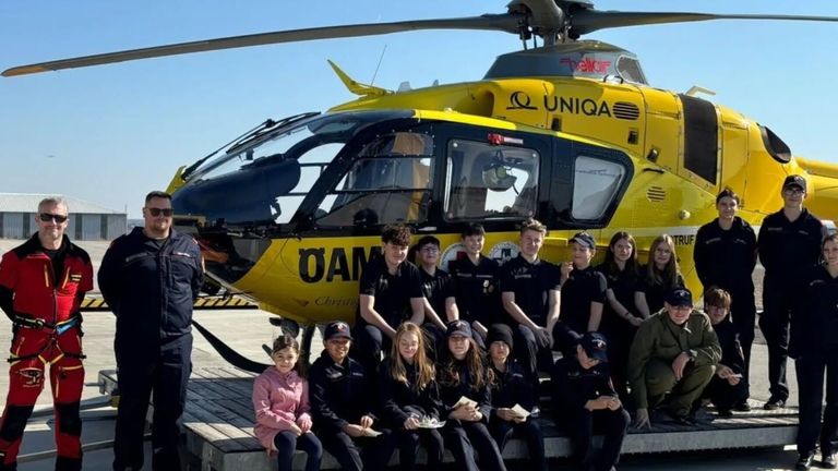 Eine Gruppe von Schülern in schwarzen Shirts und Mützen posiert vor einem gelben Bell-Hubschrauber, einige halten Zertifikate. Eine Person steht links.