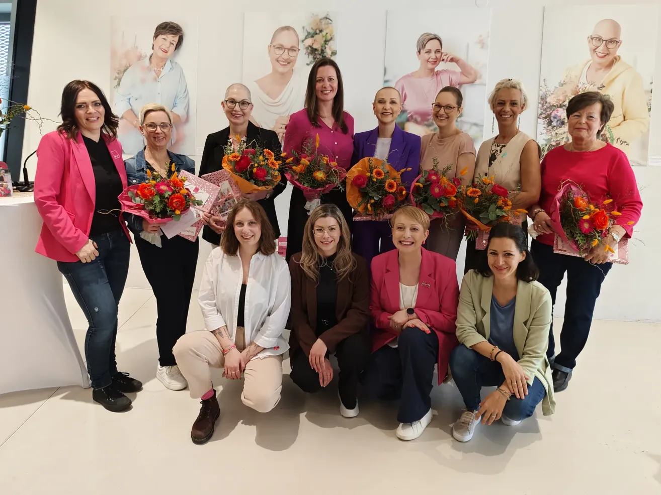 A group of women, smiling and posing for a photo, holding flowers in front of paintings on a white wall.