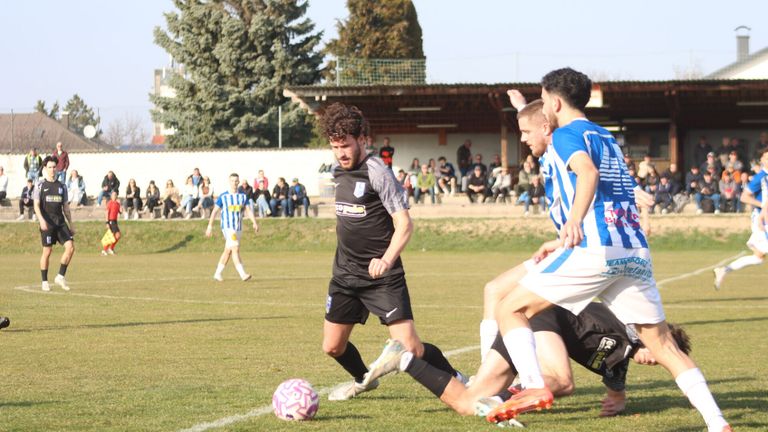 A soccer match with players on the field, one in black and white chasing a pink soccer ball, while others in blue and white defend him. Spectators watch from the bleachers.