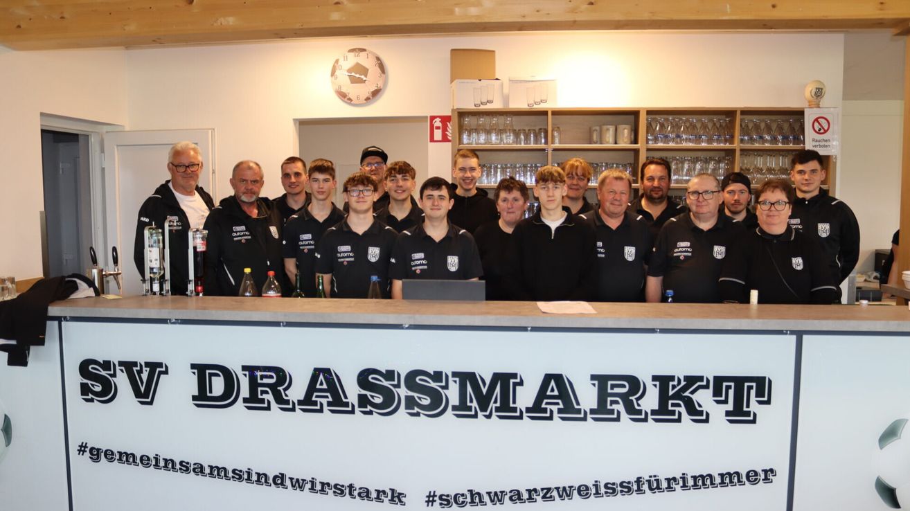 Eine Gruppe von Menschen in schwarzen Shirts steht hinter einer Theke mit der Aufschrift Drassmark, wahrscheinlich bei einer Sportveranstaltung. Einige Flaschen und ein Laptop sind auf der Theke. Ein Feuerlöscher ist an der Wand über Regalen mit Gläsern montiert.