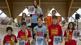 A group of young people stand together holding trophies and certificates, with one girl holding a poster. They are wearing matching jerseys and smiling for the camera.
