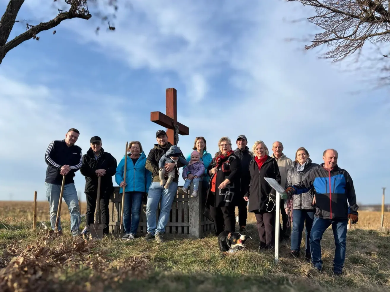 Eine Gruppe von Menschen steht auf einem Feld mit einem Holzkreuz im Hintergrund. Einige halten Schaufeln. Ein Baby wird von einem Erwachsenen in der Mitte gehalten. Ein Hund sitzt auf dem Boden.