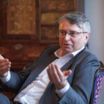 A man in a suit sits on a chair, gesturing with his hands, possibly speaking. He wears glasses and sits in front of a wooden cabinet.