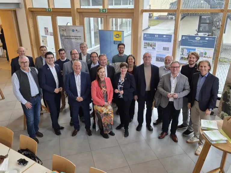 A group of professionals stands in a conference room, some wearing formal attire. Behind them, banners are displayed on the wall, and glass doors provide a view outside.
