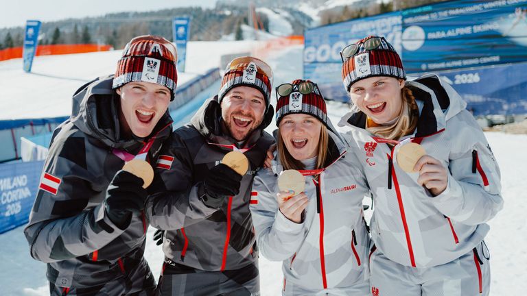 Four skiers pose for a photo with their gold medals. They are all smiling and wearing winter gear. Behind them, there is a snowy mountain and a blue banner with text.