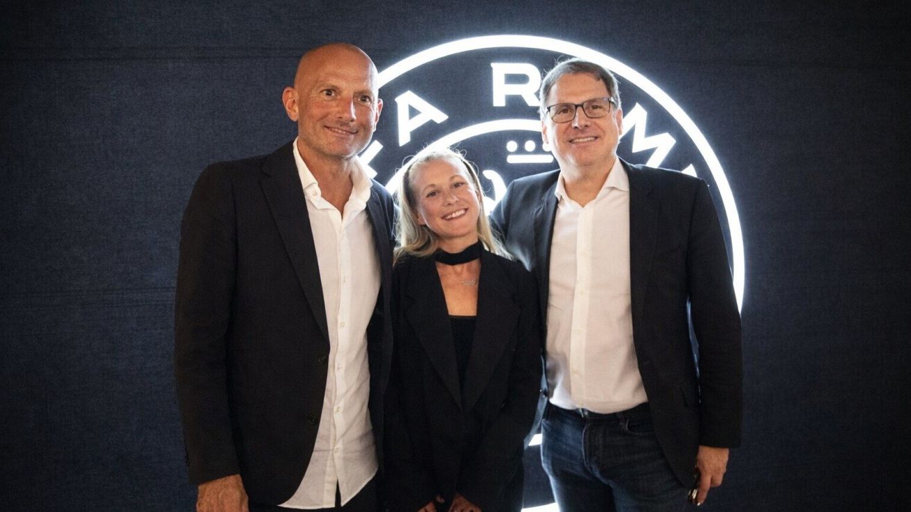 Three people stand together in front of a white logo. A man on the left, a woman in the middle, and a man on the right, all smiling. They are all wearing suits.