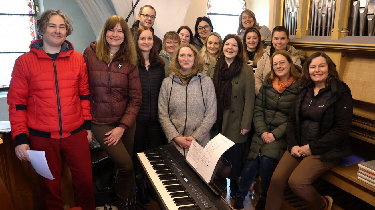 Eine Gruppe von Frauen lächelt und posiert für ein Foto, während sie in einer Kirche um ein Klavier mit Notenblättern herumstehen.