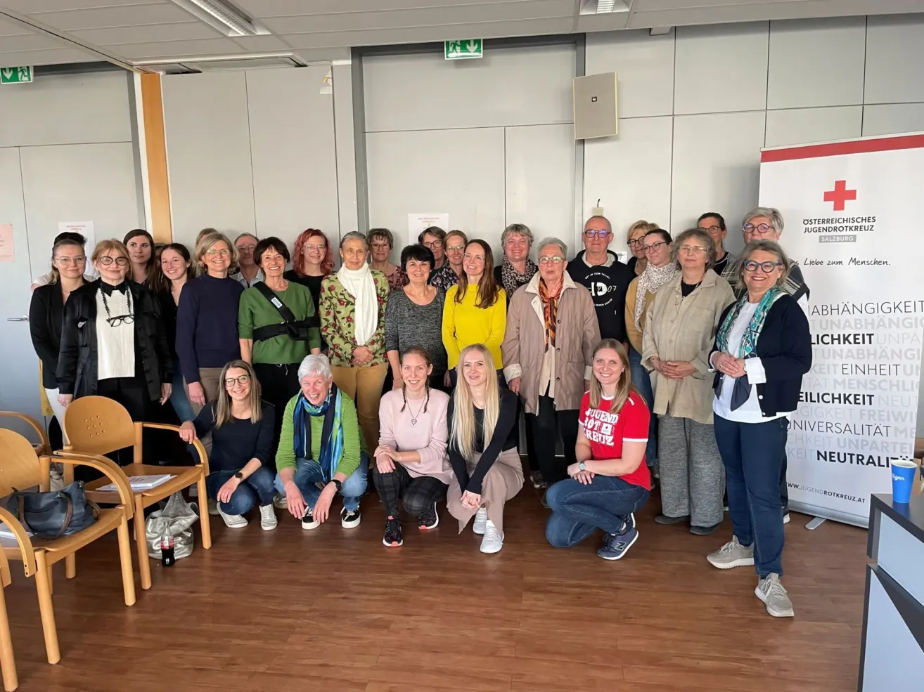 Eine Gruppe von Menschen, darunter Männer und Frauen, posiert für ein Foto in einem Raum. Einige stehen, andere sitzen auf dem Boden. Sie alle lächeln. Ein Holzstuhl steht links. Eine Tafel steht rechts.