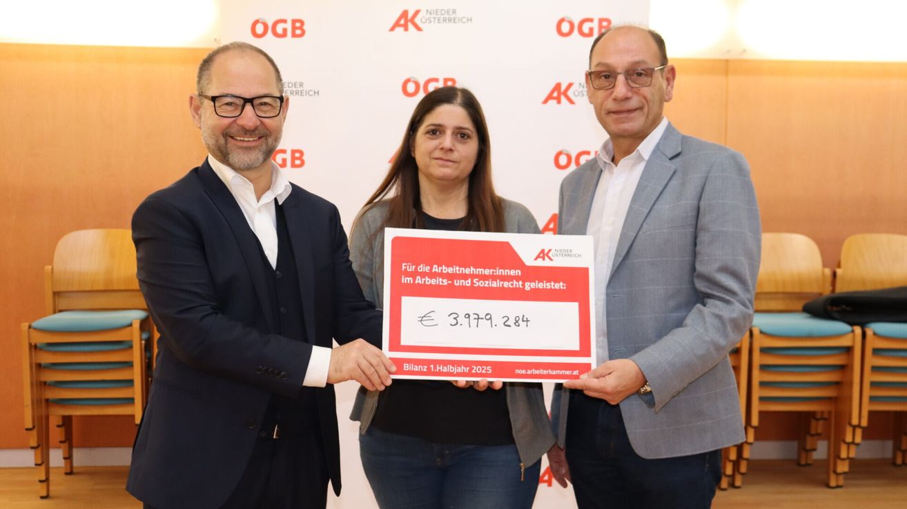 Three people stand in front of a backdrop with logos, presenting a red check for 3,979.284 Euros to a woman. The man on the left wears glasses and a suit, while the other two wear suits and eyeglasses.