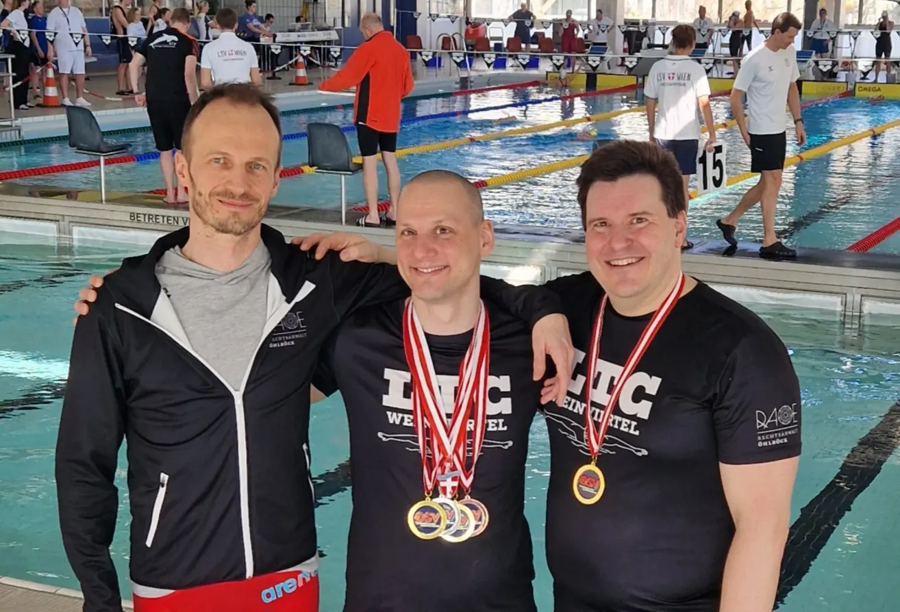 Three men wearing medals pose for a photo near a pool. One wears a black jacket, the other two black shirts. They smile for the camera.