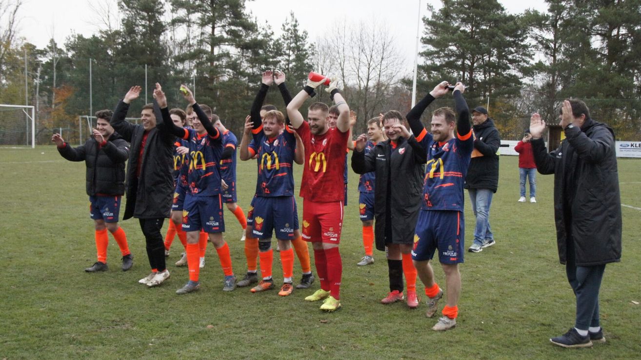 Eine Fußballmannschaft feiert einen Sieg auf einem Feld, mit Spielern in blauen und orangefarbenen Trikots, einer hält einen Ball.