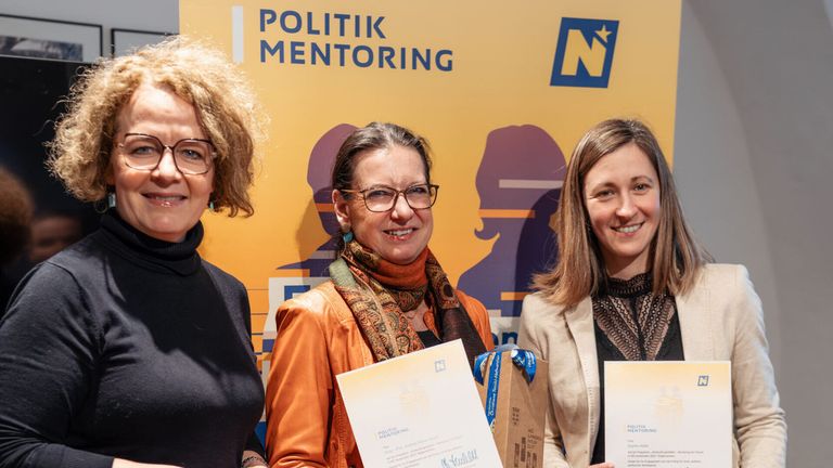 Three women hold certificates and smile for a photo in front of a yellow banner with 'POLITIK MENTORING' and an 'N' logo.