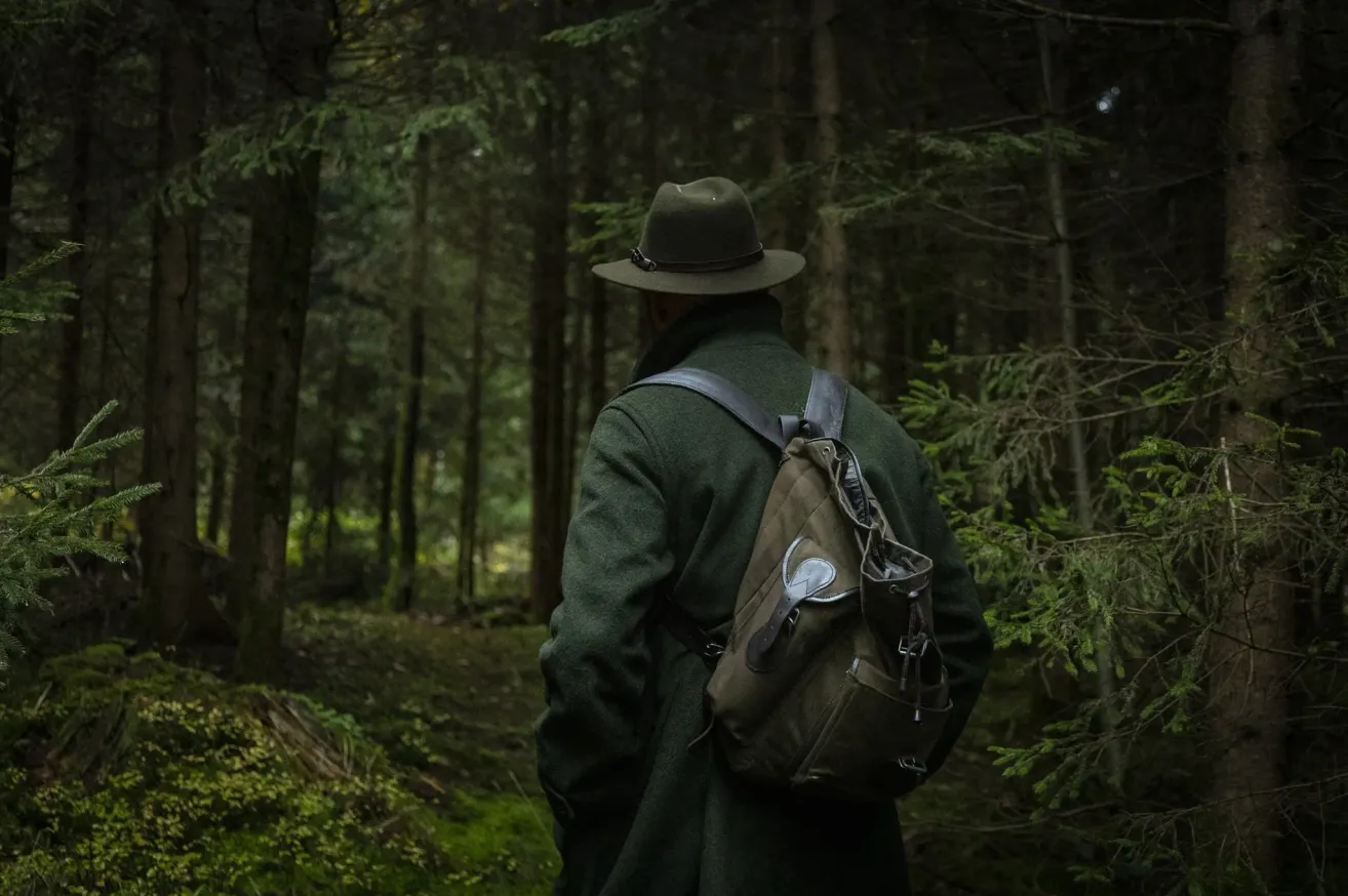 A man in a green coat and hat walks through a dense forest, carrying a green backpack.